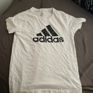 Vit Adidas t-shirt med logga - Vit t-shirt från Adidas med stor svart logga framtill. Klassisk rund halsringning och korta ärmar. Tillverkad i mjuk bomull, perfekt för en sportig och avslappnad stil.