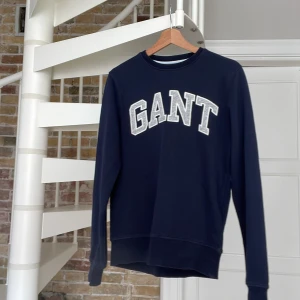 Mörkblå sweatshirt från GANT - Säljer en mörkblå sweatshirt från GANT med klassisk rund hals och stora vita bokstäver framtill. Tröjan har långa ärmar och en relaxed passform, perfekt för en chill och stilren look. Tillverkad i mjuk bomull för extra komfort.