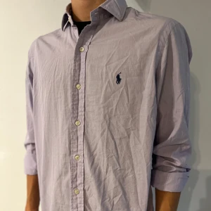 Ljuslila skjorta från Polo Ralph Lauren - Säljer en klassisk ljuslila skjorta från Polo Ralph Lauren med broderad logga på bröstet. Skjortan har lång ärm, knappar framtill och är tillverkad i bomull. Perfekt för dig som gillar stilrena och tidlösa plagg.