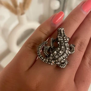 Ring med ödla i silverfärg - Cool ring i form av en ödla, täckt med glittrande stenar. Ringen är i silverfärg och har en detaljerad, tredimensionell design som verkligen sticker ut. Perfekt accessoar för dig som vill ha något unikt och edgy.