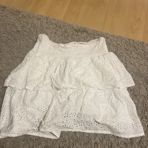 Vit volangkjol från Srn Hit - Supersöt vit kjol från Srn Hit med broderade hålmönster och flera volanger. Kjolen har smock i midjan för en bekväm passform och är gjord i bomull, perfekt för varma dagar. Dem går även att använda som en topp!