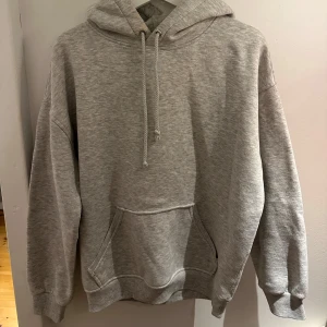 Grå hoodie med huva och magficka - Klassisk grå hoodie med huva och snörning, samt stor magficka framtill. Tröjan har en loose passform och är tillverkad i mjuk bomullsblandning. Perfekt för chill dagar eller när du vill ha en enkel och stilren look.