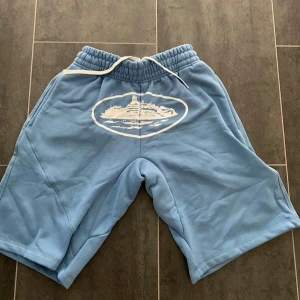 Blåa corteiz shorts - Blåa corteiz shorts med vit snörning i midjan och ett stort vitt tryck framtill. De är tillverkade i mjukt material och har en avslappnad passform. Perfekt för sommaren och vill ha sköna shorts. Hör av vid intresse, går även ner vid snabb affär. Fraktar inom 1 dag