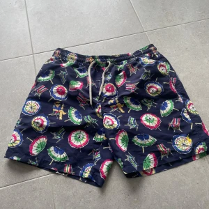 Polo Ralph Lauren badshorts - Färgglada badshorts från Polo Ralph Lauren med parasoll- och solstolsmönster på mörkblå botten. Klassisk gul broderad logga, elastisk midja med snörning och innerbyxa i mesh. Perfekta för stranden eller poolen.