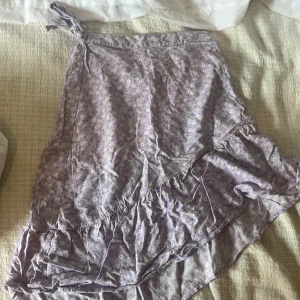 Ljuslila blommig omlottkjol Kappahl - Supersöt ljuslila omlottkjol från Kappahl med små vita blommor och volangdetalj. Kjolen knyts i sidan och har en flowy känsla, perfekt för vår och sommar. Tunn och skön bomullskvalitet som känns lätt på kroppen.