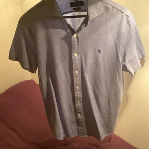 Kortärmad skjorta från Ralph Lauren - Snygg grå kortärmad skjorta från Ralph Lauren i en custom slim fit. Skjortan har knappar framtill och en liten broderad logga på bröstet. Perfekt för en stilren look.