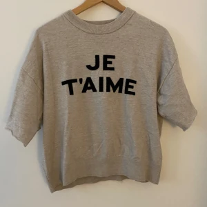 Beige tröja ifrån Gina Tricot - Snygg beige tröja från Gina Tricot med svart text 'JE T'AIME' framtill. Tröjan har korta ärmar, men när den köptes var den långarmad. Klippte den för att det passade inte med långarmad på mig. Använd och tvättad🩷🩷🩷🩷