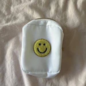 Vit liten väska med smiley-detalj - Vit liten väska med dragkedja i guld och en broderad gul smiley på framsidan. Perfekt för småprylar eller som plånbok. Enkel och clean design med en rolig detalj som sticker ut. Köpt från shein och kommer inte till användning. Kontakta gärna innan köp!