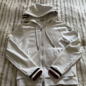 Vit Moncler kofta med ränder - Vit kofta från Moncler med dragkedja, huva och snörning. Ärmsluten och huvan har snygga ränder i mörkblå, vitt och rött. Klassisk Moncler-logga på ärmen och två fickor framtill. Perfekt för en clean och sportig look.