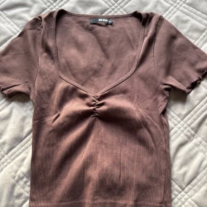 Brun ribbad topp från Bik Bok - Säljer en brun ribbad topp från Bik Bok i storlek M. Toppen har korta ärmar, djup v-ringning och snygg rynkdetalj framtill. Perfekt för dig som gillar en enkel men trendig look.