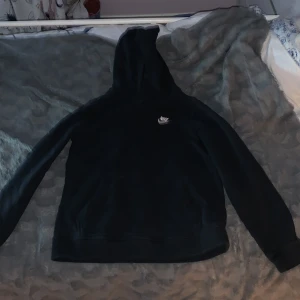 Svart Nike hoodie med broderad logga - Svart hoodie från Nike med klassisk broderad vit logga på bröstet. Tröjan har huva, lång ärm och en stor magficka framtill. Insidan är mjuk och fleecefodrad för extra komfort. Perfekt för chill dagar eller när du vill ha en sportig look.