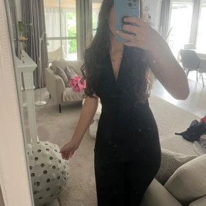 Svart jumpsuit med v-ringning är uppen rygg där bak  - Snygg svart jumpsuit med djup v-ringning och markerad midja. Byxorna är långa och vida, vilket ger en stilren och modern look. Tillverkad i stretchigt material. Perfekt för dig som vill sticka ut med en enkel men elegant outfit.🌸