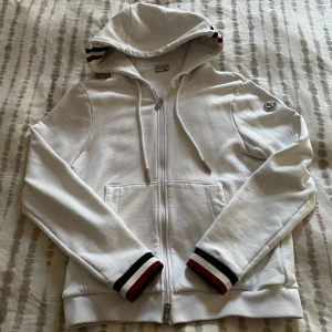 Vit kofta från Moncler med ränder - Snygg vit kofta från Moncler med dragkedja, huva och snörning. Ärmsluten och huvan har stilrena ränder i mörkblå , vitt och rött. Klassisk Moncler-logga på ärmen och två fickor framtill. Perfekt för en clean och sportig look.