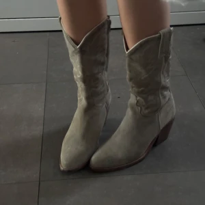 Beiga mocka cowboy boots - Snygga cowboy boots i beige mocka med spetsig tå och låg klack. Klassisk western-design med dekorativa sömmar och högt skaft. Perfekta för dig som vill sticka ut med en cool och trendig look.