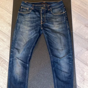 Blå jeans från Nudie Jeans Co - Säljer ett par snygga blå jeans från Nudie Jeans Co med klassisk orange söm och cool slitning framtill. Jeansen har raka ben och fem fickor, samt Nudie-logga på bakfickan. Perfekta för dig som gillar en avslappnad men stilren look. De är storlek W31-L32. Sitter slimfit och har en bra passform. Skriv till mig om du har några funderingar!😆