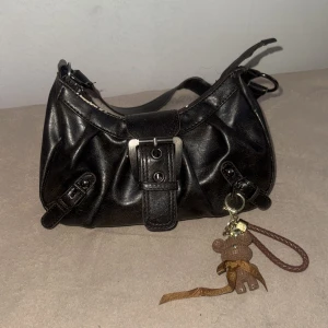 Svart axelväska med nalleberlock - Cool svart axelväska i skinnimitation med snygga spänndetaljer och dragkedja upptill. Väskan har en bred axelrem och en söt brun nalleberlock med rosett som hänger på sidan. Perfekt storlek för det viktigaste och ger en edgy vibe till din outfit.