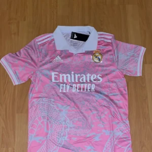  rosa fotbollströja   - Säljer en rosa Real Madrid fotbollströja från Adidas i storlek S. Tröjan har korta ärmar, vit krage och ett unikt mönster med drakar i ljusgrått. Klubbmärke och Adidas-logga på bröstet samt 'Emirates Fly Better' tryckt framtill. Tillverkad i lätt funktionsmaterial.
