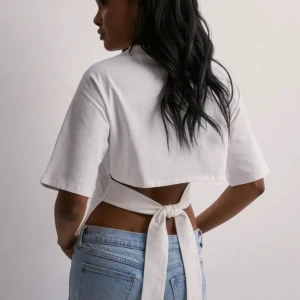 Open back tie tee, knyttröja - Säljer en vit croppad topp med korta ärmar och snygg knytning i ryggen. Toppen har en enkel och clean look framtill, men baksidan har öppen rygg och breda knytband som ger en cool twist. Perfekt till jeans eller kjol för en trendig vibe.