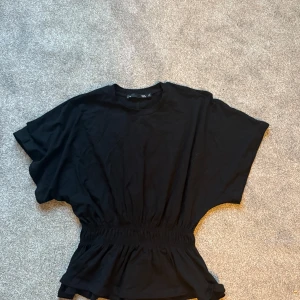 Svart topp lager 157 - Svart topp från lager 157 i mjuk bomull med bred resår i midjan som ger en snygg. Modellen har korta ärmar och rund halsringning. Perfekt för dig som gillar en enkel men ändå cool look.