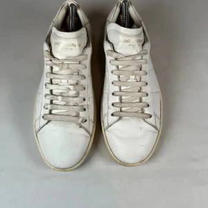 Vita Saint Laurent SL/01 sneakers - Klassiska vita Saint Laurent SL/01 sneakers i skinn med rund tå och platt sula. Diskret guldtext på plösen och stilrena paneler på sidorna. Tjocka vita snören och en minimalistisk vibe som passar till allt. Perfekta för dig som gillar lyxiga och cleana sneakers. Kommer utan box o og 100% äkta har bevis