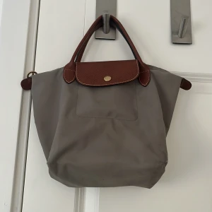 Grå Longchamp handväska med skinn - Säljer en klassisk grå handväska från Longchamp med bruna skinnhandtag och lock. Väskan har guldknapp med logga och är tillverkad i slitstark syntet med detaljer i skinn. Perfekt storlek för vardagsgrejer och enkel att bära i handen. Vet inte exakt stolek men tror S. Pris kan diskuteras