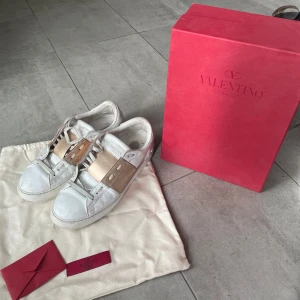 Valentino Garavani Open sneakers vit/guld - Snygga Valentino Garavani Open sneakers i vitt skinn med bred metallic guldrem över vristen och coola nitar baktill. Klassisk rund tå, platt sula och snörning.  Skorna är vält andvända och defekterna ser ni på bilderna därav det lägre priset. Ifall ni vill ha närmare bilder på skorna är det bara till att fråga❣️