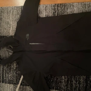Svart vindjacka från The North Face - Svart vindjacka från The North Face med huva och dragkedja framtill. Jackan har en clean look med diskret logga på bröstet och en praktisk bröstficka med dragkedja. Perfekt för blåsiga dagar och enkel att matcha med allt.