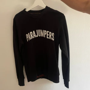 Svart långärmad tröja Parajumpers - Svart långärmad tröja från Parajumpers med vit logga tryckt över bröstet och ett broderat märke på ärmen. Tröjan har rund halsringning och är tillverkad i mjuk bomull, perfekt för en avslappnad och stilren look.
