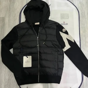 Svart cardigan med huva från Moncler - Snygg svart pufferjacka från moncler med huva och dragkedja. Jackan har stickade ärmar med vit detalj och moncler-logga på ena ärmen, perfekt inför hösten och kyligare dagar, passar mig som e 184 perfekt