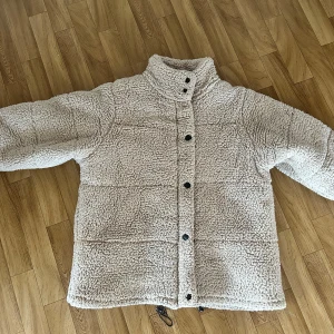 Beige teddyjacka från Boohoo - Supermysig beige teddyjacka från Boohoo med hög krage och svarta knappar framtill. Jackan har ett fluffigt yttre i teddy och slätt ljust foder på insidan. Perfekt för kalla dagar när du vill vara både varm och snygg.