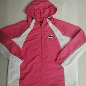 Cortiez Rosa och vit vindjacka med huva - Snygg rosa vindjacka med vita detaljer och huva. Jackan har dragkedja framtill, elastiska muddar och en cool logga på bröstet. Perfekt för dig som gillar sportig stil och vill sticka ut med färg. Passar till både chill och aktiva dagar.