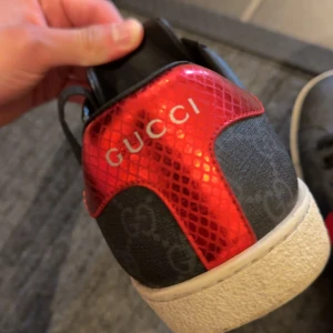 Gucci sneakers med GG-mönster och röd detalj - Snygga svarta Gucci sneakers med GG-monogram över hela skon, klassiska röda och marinblå ränder på sidorna och metallicröd samt mörkblå hälkappa med ormskinnsmönster. Skorna har svarta snören och vit sula. Perfekt för dig som vill sticka ut med lyxig streetstyle.