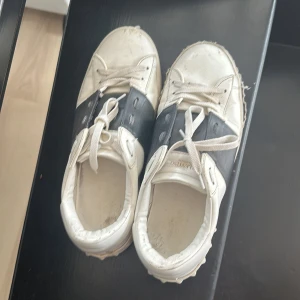 Valentino Open sneakers vit/svart - Snygga Valentino Open sneakers i vitt skinn med bred svart läderdetalj över mitten. Klassisk rund tå, vita snören och coola nitar längs sulan. Perfekta för dig som gillar streetstyle med lyxig touch.