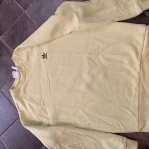 Gul sweatshirt från Adidas Originals - Gul sweatshirt från Adidas Originals med broderad svart logga på bröstet. Tröjan har rund halsringning och långa ärmar. Tillverkad i mjuk bomullsmix, perfekt för en chill och sportig stil.