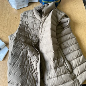 Beige dunväst från Massimo Dutti - Snygg beige dunväst från Massimo Dutti med quiltad design och hög krage. Västen har dragkedja framtill och en stilren knappdetalj vid halsen. Perfekt att slänga över en hoodie eller tröja för extra värme och stil.