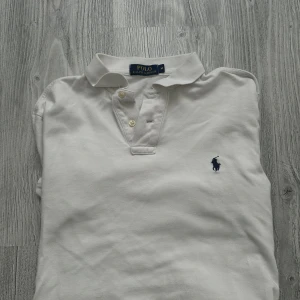 Vit långärmad pikétröja Ralph Lauren - Vit långärmad pikétröja från Polo Ralph Lauren med klassisk krage och knäppning framtill. Tröjan har den ikoniska blå broderade loggan på bröstet och är tillverkad i bomull för en skön och stilren look. Har ej använt den mycket den är lite för liten på mig 