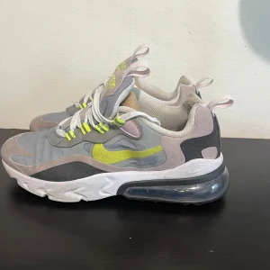 Nike Air Max 270 React sneakers grå/gul - Nike Air Max 270 React sneakers i grått, svart och beige med detaljer i neon-gult. Ovandel i textil och syntet, vit sula med synlig Air Max-bubbla. Skorna har snörning och coola kontrastdetaljer. Perfekta för dig som gillar streetstyle och sportig vibe.