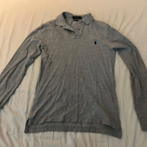 Grå långärmad pikétröja Polo Ralph Lauren - Säljer en grå långärmad pikétröja från Polo Ralph Lauren i slim fit. Tröjan har klassisk krage, knäppning framtill och den ikoniska broderade loggan på bröstet. Perfekt för dig som gillar stilrena och tidlösa plagg. Mycket bra skick bara lite skrynklig 