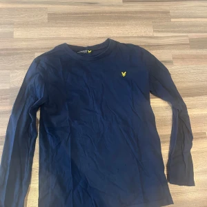 Mörkblå långärmad tröja Lyle & Scott - Snygg mörkblå långärmad tröja från Lyle & Scott med klassisk gul logga på bröstet. Tröjan är i mjuk bomull och har en enkel, stilren design som passar perfekt till jeans eller chinos.