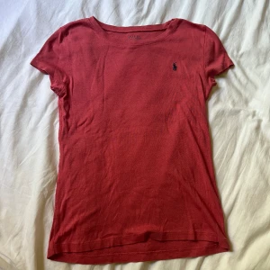 Röd t-shirt från Polo Ralph Lauren - Röd t-shirt från Polo Ralph Lauren med klassisk rund hals och korta ärmar. T-shirten har en liten mörkblå broderad logga och är gjord i mjuk bomull. 