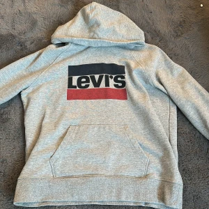 Grå Levi's hoodie med logga, M - Snygg grå hoodie från Levi's i bomull med stor logga i svart, vitt och rött framtill. Klassisk känguruficka, huva och broderad detalj på ärmen. Perfekt för chill dagar och streetstyle.