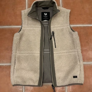 Beige fleeceväst från Ragwear - Mysig beige fleeceväst från Ragwear i storlek S. Västen har hög krage, hel dragkedja framtill och en bröstficka med dragkedja. Perfekt för lager-på-lager-stil.