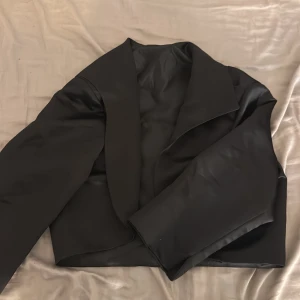 Svart croppad kavaj i satin-look - Snygg svart croppad kavaj med breda slag och lång ärm. Jackan har en stilren och minimalistisk design med en lätt glansig satin-finish som ger en lyxig känsla. Perfekt för dig som vill sticka ut med en modern och edgy look.