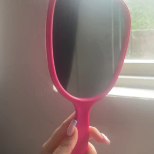 Handhållen sminkspegel i rosa - Snygg handhållen sminkspegel med starkt rosa ram och handtag i plast. Baksidan har ett mönster med vågformade cirklar för extra grepp. Perfekt storlek för att fixa sminket snabbt och enkelt. Spegeln har ett hål i handtaget för enkel upphängning.