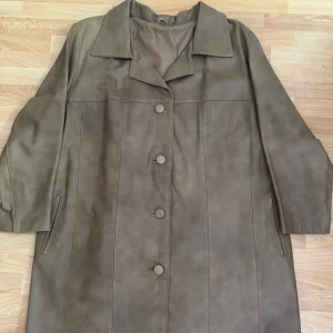 Beige skinn rock med knappar - Snygg beige rock i mjuk skinn/äkta skinn/ med klassisk krage och stora matchande knappar framtill. Jackan har raka linjer, diskreta fickor i sidorna och lång ärm. Perfekt för dig som gillar stilren och tidlös look. Använd en gång. Sydde på beställning, längd 92cm, bröst 124cm, ärmar 56cm.