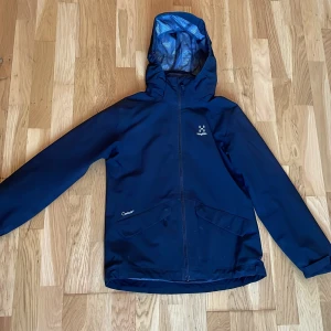Svart vindjacka från Haglöfs - Snygg mörkblå vindjacka från Haglöfs med huva och meshfoder. Jackan har dragkedja framtill, två fickor och justerbara ärmslut. Använt en gång 