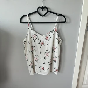 Vit blommig linnetopp med smala axelband - Supersöt vitt linne med tunna axelband och blommigt mönster i rosa och grönt. Toppen har en avslappnad passform och lätt v-ringad halsringning. Toppen ger en lätt och luftig känsla, passar till både jeans och kjol.