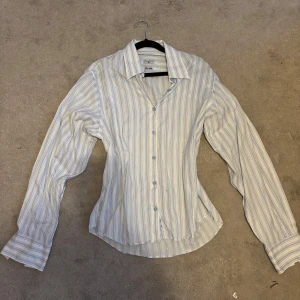 Randig skjorta från Buxbom XL (passar mer som en M) - Klassisk skjorta från Buxbom i vitt med tunna blå ränder. Skjortan har lång ärm, knappar framtill och en snygg krage. Tillverkad i bomull och har en normal passform. Perfekt för dig som gillar stilrena och fräscha plagg.