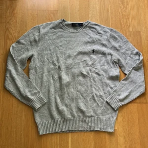 Grå stickad tröja från Polo Ralph Lauren - Snygg grå stickad tröja från Polo Ralph Lauren med klassisk rund halsringning och diskret broderad logga på bröstet. Tröjan har ribbade muddar vid ärmslut och nederkant, perfekt för lager-på-lager under kyliga dagar.