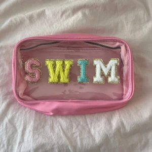 Rosa necessär med SWIM-bokstäver - Söt rosa necessär med transparent framsida och färgglada bokstäver som stavar SWIM. Väskan har dragkedja i guld och är perfekt för att förvara småsaker till stranden eller smink. Den är köpt från shein och kommer inte mer till användning. Kontakta gärna innan köp!